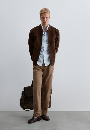 Homme portant une veste marron, une chemise bleu clair, un pantalon beige et des chaussures marron foncé, debout avec les mains derrière le dos et un sac foncé sur le sol.