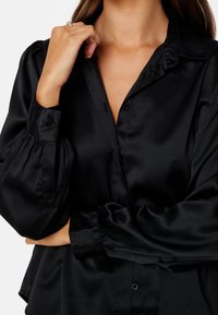 Blusa nera in satin con chiusura a bottoni frontale, colletto e maniche lunghe a sbuffo con polsini arricciati. Texture liscia e finitura lucida.