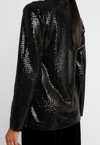 Černý flitrový blejzr s texturovaným povrchem, s volným střihem a dlouhými rukávy. Lesklé flitry vytvářejí odrazový efekt.