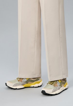 Osoba w beżowych, szerokich spodniach i wielokolorowych sneakersach z żółtymi, zielonymi, beżowymi i białymi akcentami, stojąca na neutralnym tle.