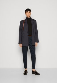Navyblazer im entspannten Schnitt, kombiniert mit einem schwarzen Rollkragenpullover und dunklen Hosen. Abgerundet mit schwarzen Schuhen und einer braunen Schultertasche.