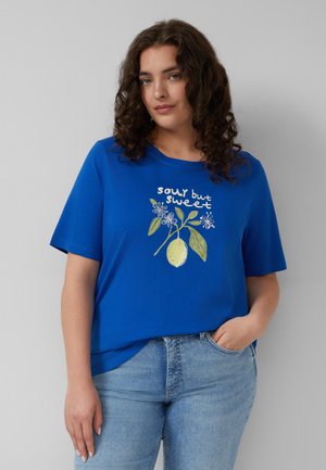 Femme aux cheveux bouclés portant un t-shirt bleu avec une illustration de citron et de fleurs et le texte « acidulé mais doux » associé à un jean bleu clair.