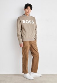 Sudadera de color marrón claro con "BOSS" impreso en blanco, combinada con pantalones cargo beige y zapatillas blancas. El modelo está de pie contra un fondo neutro.