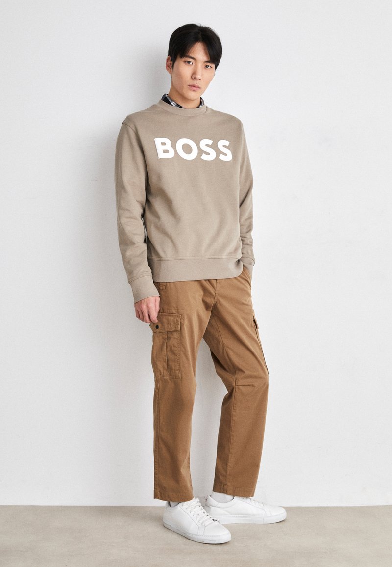 Felpa beige con la scritta 'BOSS' in bianco, abbinata a pantaloni cargo color sabbia e sneakers bianche. Il modello è in piedi davanti a uno sfondo neutro.
