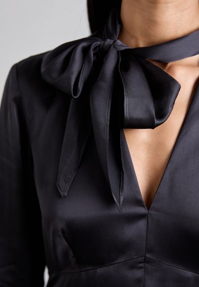 Schwarze Satinbluse mit einem auffälligen Schleifenkragen, V-förmigem Ausschnitt und glatter Textur, die das elegante Design und die Details hervorhebt.