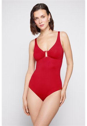 Žena s krátkými tmavými vlasy, oblečená v jednobarevných červených jednodílných plavkách s obdélníkovým výřezem na hrudi, pózující před bílým pozadím.