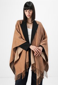 Brun mønster poncho med sort blazer-lignende krave, frynsekant og tekstureret stof. Båret over en hvid top med denimbukser.
