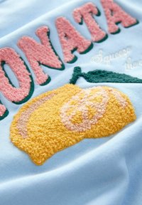 Blauer Stoff mit rosa strukturierten Stickereien, die "Sonata" buchstabieren, sowie einem gelben, flauschigen Zitronendesign und grünen Blättern, die Tiefe und Farbe hinzufügen.