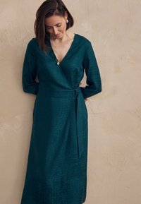 Femme en robe portefeuille teal à manches longues, debout contre un mur beige texturé, regardant vers le bas avec les mains derrière le dos.