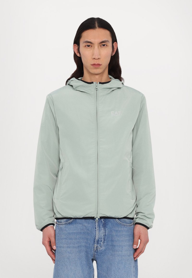 Lys mintgrøn zip-up jakke lavet af letvægtsmateriale med hætte, der har et frontlogo, elastiske ærmer og to sidelommer.