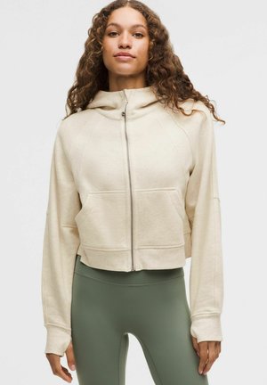 Žena so kučeravými vlasmi, oblečená v béžovej mikine so zapínaním na zips a olivovo zelených legínach s vysokým pásom, na jednoduchom bielom pozadí.