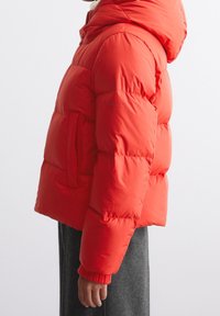 Czerwona kurtka puchowa z kapturem, krótki fason, detal w postaci ściśniętych mankietów oraz kieszony bocznej. Tkanina wydaje się błyszcząca i pikowana, zapewniając izolację.