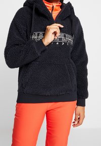 Schwarzer Kapuzenpullover aus Fleece mit strukturiertem Stoff, Kängurutasche und weißem Logo-Print auf der Brust. Kombiniert mit leuchtend orangefarbener Hose.