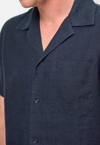 Camicia a maniche corte di colore blu navy realizzata in tessuto testurizzato, con colletto a punta, taschino sul petto e bottoni scuri lungo la parte anteriore.