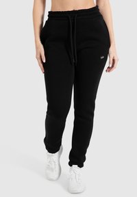 Schwarze Fleece-Jogginghosen mit elastischem Bund, verstellbarem Kordelzug, Seitentaschen und einem kleinen Logo-Detail am Oberschenkel. Weiße Sneaker.