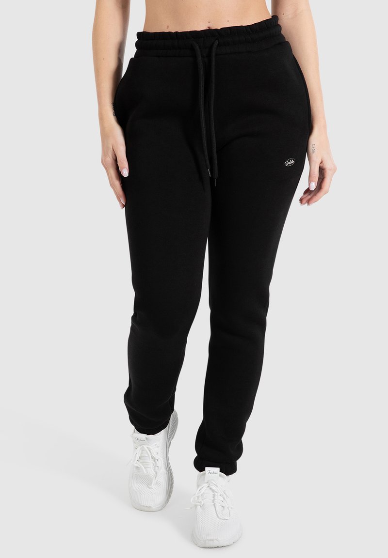 Schwarze Fleece-Jogginghosen mit elastischem Bund, verstellbarem Kordelzug, Seitentaschen und einem kleinen Logo-Detail am Oberschenkel. Weiße Sneaker.