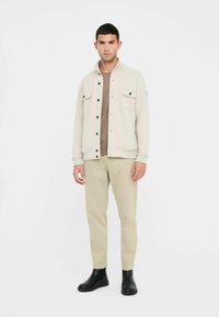 Giacca grigio chiaro con due tasche frontali, bottoni neri; indossata sopra una camicia marrone e abbinata a pantaloni beige e stivaletti neri.