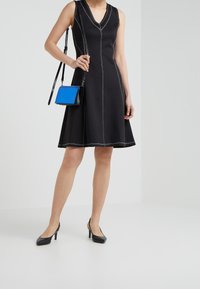 Vestito nero senza maniche con cuciture bianche, orlo svasato; abbinato a una piccola borsa a tracolla blu e scarpe con tacco alto nere.