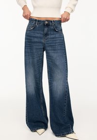 Wide-leg blue denim jeans with a slightly faded finish, five-pocket design, and a button closure at the waist. Soft, textured fabric.