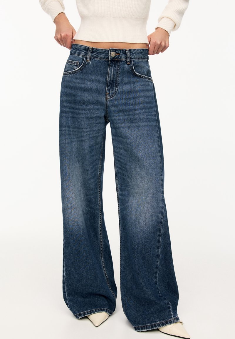 Wide-leg blue denim jeans with a slightly faded finish, five-pocket design, and a button closure at the waist. Soft, textured fabric.