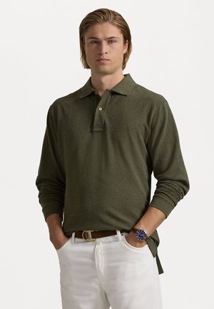 Olijfgroene longsleeve polo met kraag, met een tonale logo. Draagt met witte broeken en een bruine riem, met een blauwe horloge.