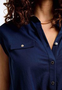 Personne aux cheveux bouclés portant une chemise sans manches bleu marine à boutons avec une poche poitrine et un fin collier en or.