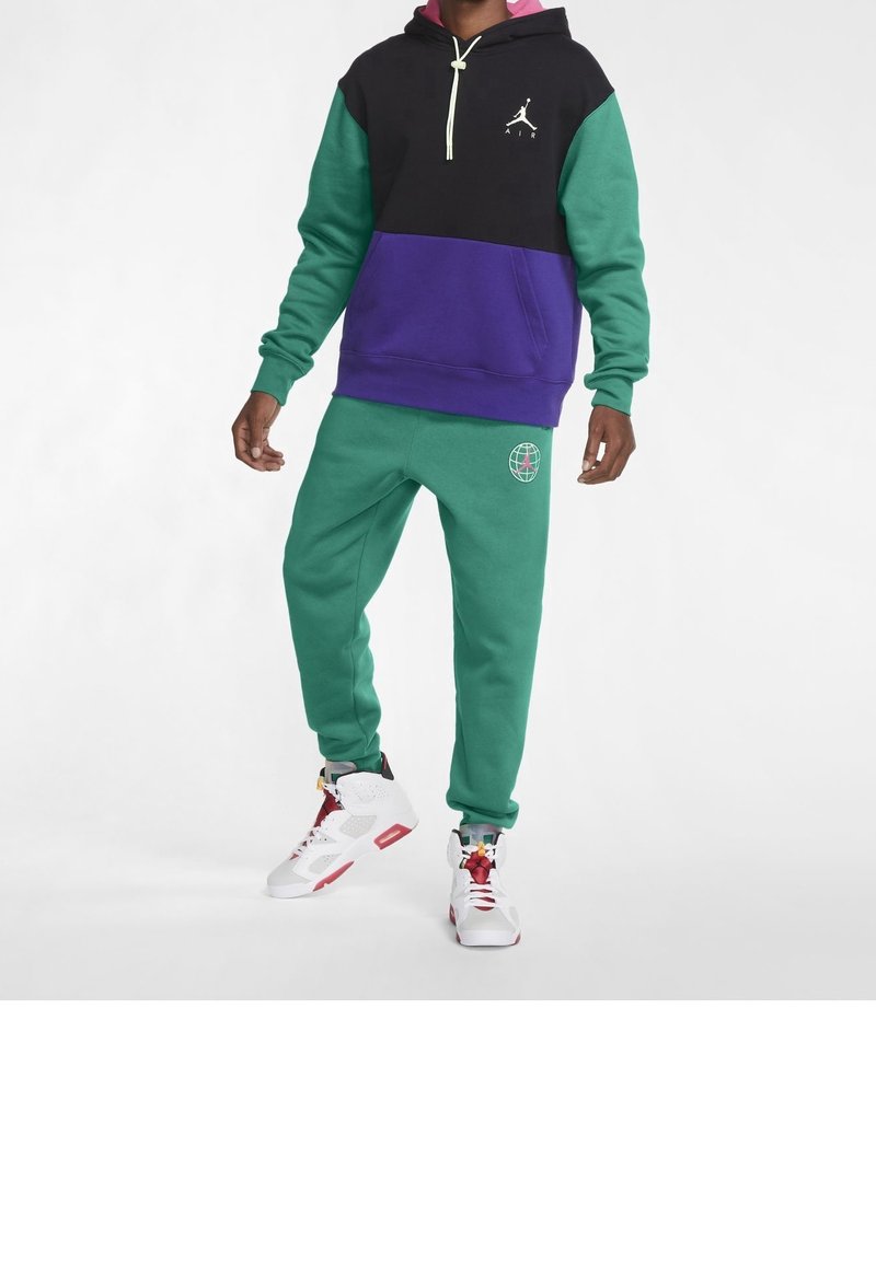 Person klädd i en svart, grön och lila Jordan-hoodie med gröna sweatpants och vita och röda Jordan-sneakers stående på vit bakgrund.