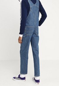 Pantalones de mezclilla en azul medio con tirantes ajustables, dos bolsillos delanteros y un corte recto. Combinados con zapatillas moradas y calcetines blancos.