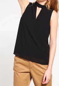 Top nero senza maniche con colletto alto e scollatura a chiave, caratterizzato da un orlo inferiore trasparente e tessuto morbido. Indossato con pantaloni beige chiaro.