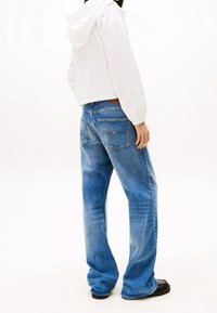 Persoon die losse blauwe spijkerbroek, een witte kapotte jas en zwarte schoenen draagt, staande tegen een effen lichte achtergrond met de rug naar voren.