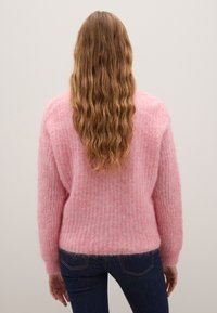Pullover rosa, soffice e dal taglio morbido, con texture a coste, spalle scese e maniche lunghe, indossato sopra jeans scuri.