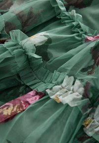 Tessuto verde trasparente con stampa floreale e bordi arricciati, che mostra delicati dettagli in rete arricciati e una morbida consistenza.