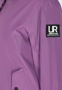Veste imperméable violette avec poche latérale et écusson noir portant l'inscription « UR Urban Rain » sur la manche supérieure.