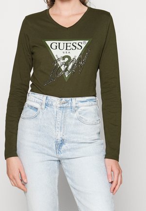 Vrouw draagt een olijfgroen shirt met lange mouwen en Guess-logo en lichtblauwe hooggetailleerde jeans, staand tegen een effen achtergrond.