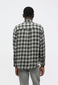 Άντρας γυρισμένος με πλάτη, φοράει μαύρη και άσπρη καρό πουκαμίσα με μακριά μανίκια και ανοιχτό γκρι τζιν, σε έναν απλό λευκό φόντο.