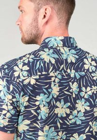 Camisa de manga corta con fondo azul marino, que presenta grandes patrones florales en azul claro y crema, textura tejida y cuello abotonado.
