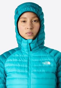 Tyrkis pufferjacka med huva, med quiltad textur, hög krage och dragkedja framtill. Logotyp på vänster bröst, lättviktsdesign.