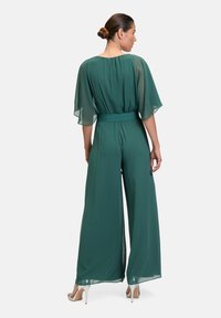 Jumpsuit verde smeraldo in chiffon leggero, con maniche a volant, vita con cordoncino e design a gamba larga. Texture morbida e silhouette fluida.