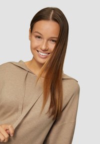 Beige Kapuzenpullover mit einem lässigen Schnitt, ausgestattet mit einer Kapuze, einem dezenten V-Ausschnitt und weichem Material. Das Modell hat lange, glatte Haare.