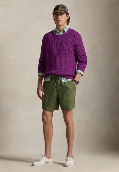 Μωβ πουλόβερ με πλέξη καλτσοδέτα πάνω από πράσινο σορτς με κορδόνι. Το ντύσιμο περιλαμβάνει μια ριγέ μπλούζα από κάτω και λευκά παπούτσια με κορδόνια.