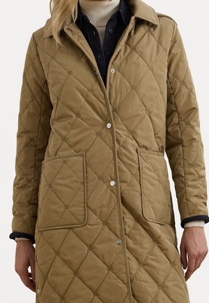 Femme portant un long manteau matelassé marron avec de grandes poches avant, par-dessus une chemise noire et un pull à col roulé beige.