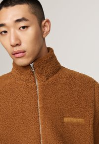 Brun plysj zip-up jakke med høy krage og teksturert stoff. Har en sølvfarget glidelås og et brunt logo-patch på brystet.
