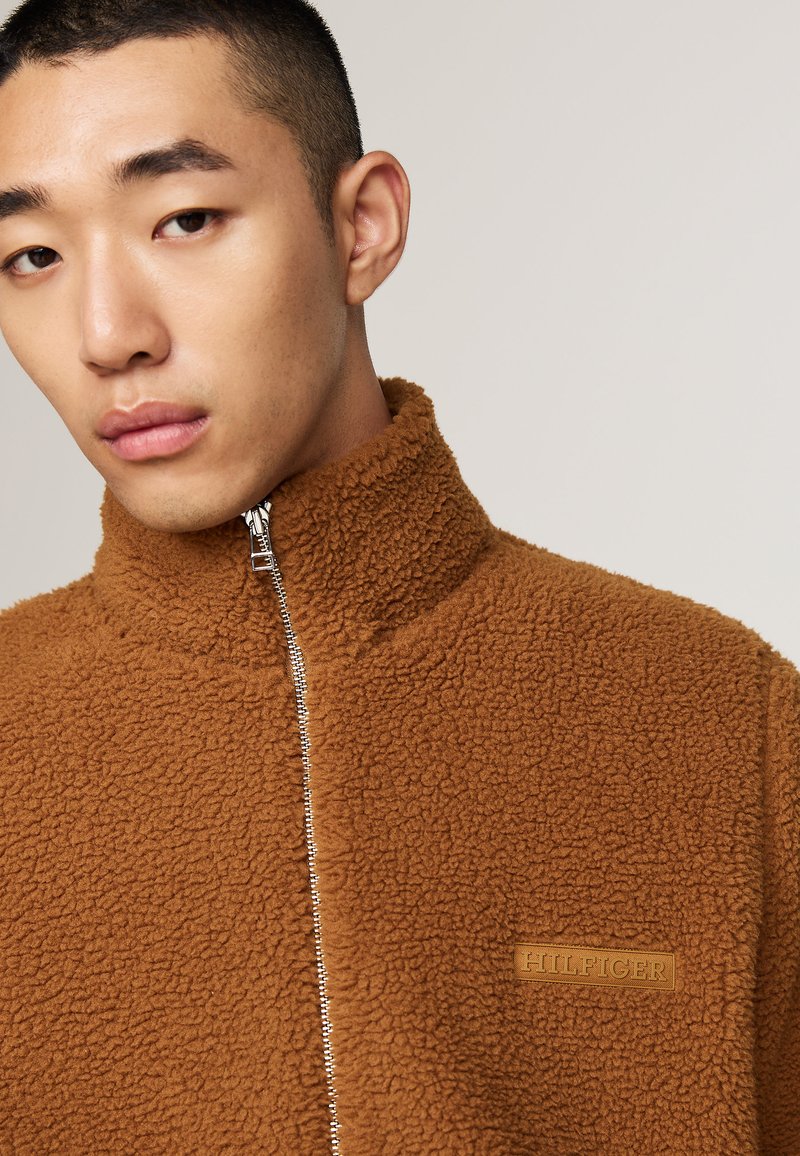 Giacca in pile marrone con zip e collo alto, realizzata in un tessuto testurizzato. Presenta una zip argentata e un patch con logo marrone sul petto.