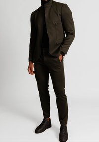 Completo color verde oliva scuro con un design aderente. La giacca presenta un colletto alto e una chiusura con zip. Abbinato a pantaloni coordinati e sneakers nere.