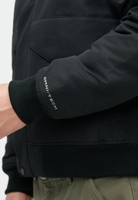 Μαύρο παλτό puff με ριμπ μανσέτα, φερμουάρ στις τσέπες και ετικέτα "OMNI-TECH" στο μανίκι. Λείο, μονωμένο ύφασμα.