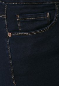 Donkerdenim jeans met een zichtbare voorzak, koperen knop en contrasterende beige stiksels langs de randen. Glad textuur.