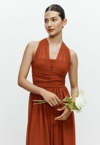 Abito color ruggine con scollo all'americana, tessuto testurizzato, vita arricciata e gonna fluente. La modella tiene una rosa bianca e indossa un anello.