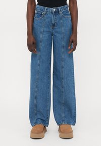 Person med høy midje, vide blå jeans med front sømdetaljer og brune semskede platåsko mot en enkel bakgrunn.