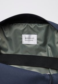 Laavinsininen laukku, jossa on pehmeä pinta, vetoketjusulku ja vihreä sisävuori; tarrassa lukee "valmistettu luomupuuvillasta ja kierrätetystä polyesteristä."