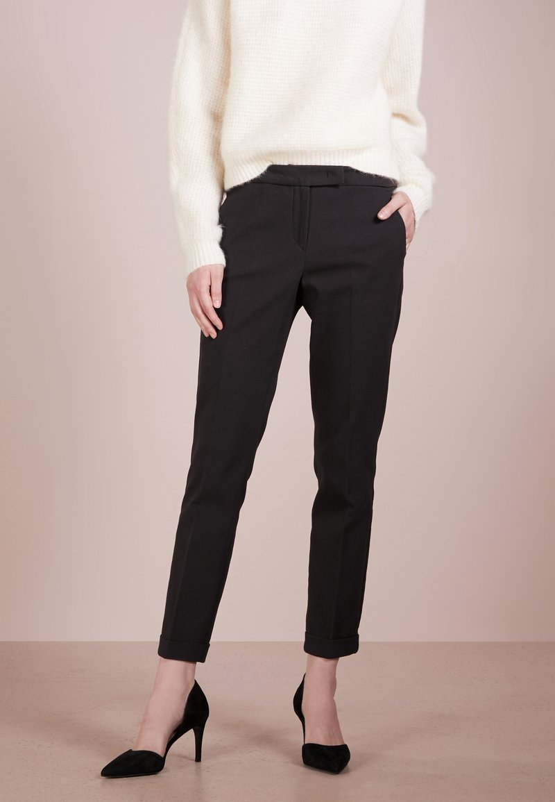 Femme portant un pantalon noir coupé sur mesure, longueur cheville avec des revers, associé à des talons hauts noirs et un pull blanc duveteux, debout sur un sol beige.